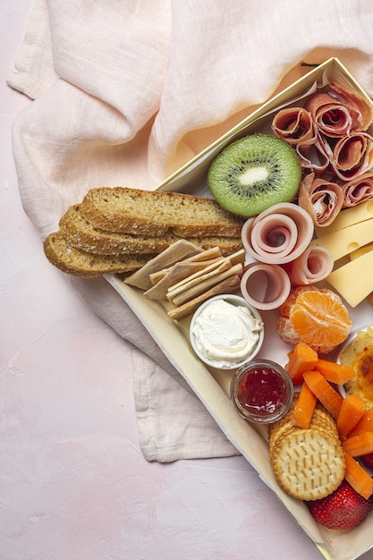 Gesunde Brunchbox zum Mitnehmen mit Schinken, Erdbeeren; Kiwi; Brot; Kekse; Käse, Karotten, Mandarinen, Hummus.