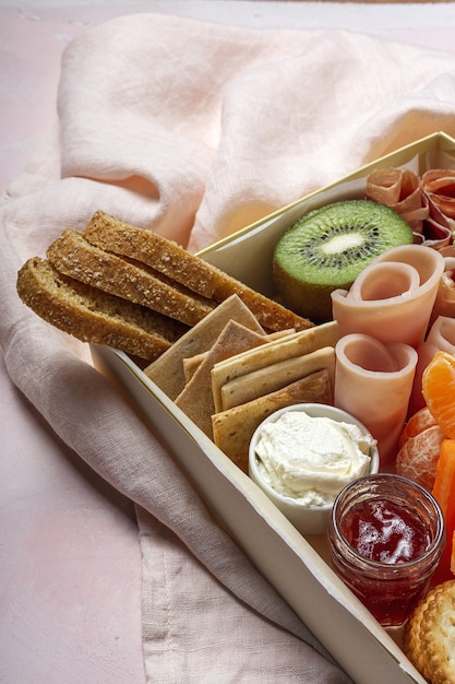 Gesunde Brunchbox zum Mitnehmen mit Schinken, Erdbeeren; Kiwi; Brot; Kekse; Käse, Karotten, Mandarinen, Hummus.