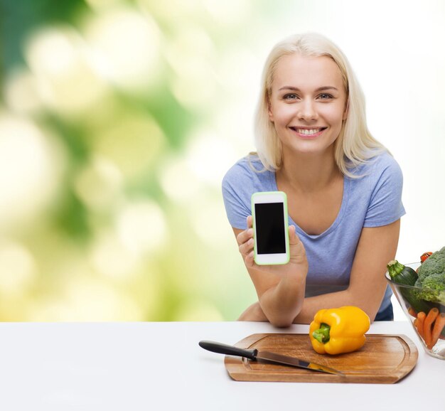 Gesunde Ernährung, vegetarisches Essen, Diäten und People-Konzept - lächelnde junge Frau, die Gemüse kocht und einen leeren Smartphone-Bildschirm auf grünem, natürlichem Hintergrund zeigt