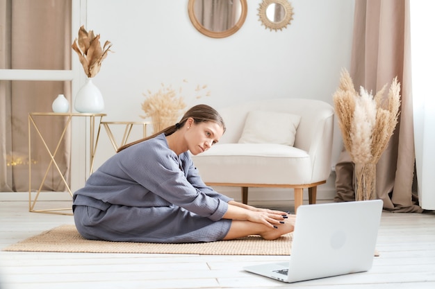 Gesunde Frau, die sich Videos auf dem Laptop ansieht, während sie zu Hause auf der Yogamatte trainiert