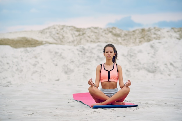 Gesunde Frau, die Yoga praktiziert und am Strand im Sand meditiert