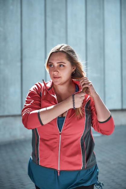 Gesunde Frau. Vertikales Foto einer hübschen Frau in Übergröße in Sportkleidung, die ihr langes Haar anpasst und wegschaut, während sie im Freien steht