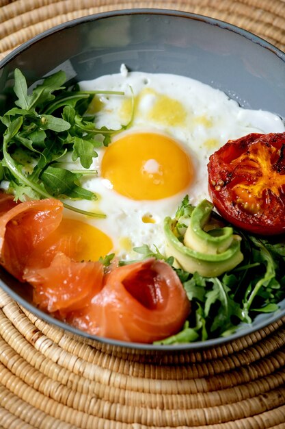 Gesunde Frühstücksschüssel mit Spiegeleiern, Lachs, Avocado, gegrillter Tomate und Salat, serviert mit Brot auf Strohserviette