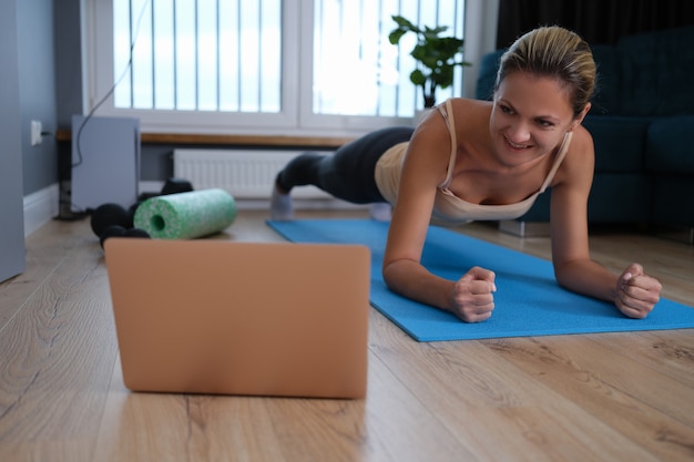 Gesunde junge Frau, die zu Hause Plankenübungen macht, während sie sich eine Online-Trainingssitzung vom Laptop aus ansieht