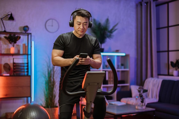 Gesunde Passform lächelnder asiatischer Mann, der zu Hause auf einem statischen Heimtrainer während des Trainings mit Telefon trainiert