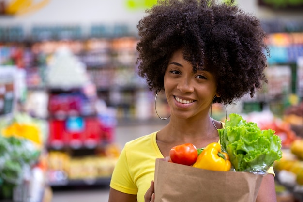 Gesunde positive glückliche afrikanische Frau, die eine Papiereinkaufstasche voll von Obst und Gemüse hält.