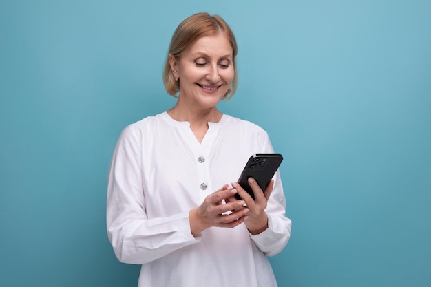 Gesunde, schlanke, blonde, reife Frau, die die Technik mit dem Smartphone auf Studiohintergrund beherrscht