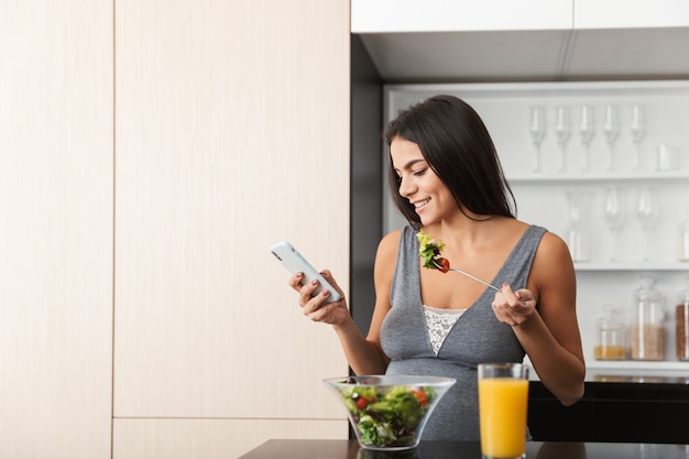 Gesunde schwangere Frau drinnen zu Hause in der Küche mit Handy essen Salat.