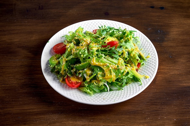 Gesunde und diätetische Salatmischung mit Avocado, Sesam, gelbem Dressing und Kirschtomaten, serviert in einem weißen Teller auf einer Holzoberfläche