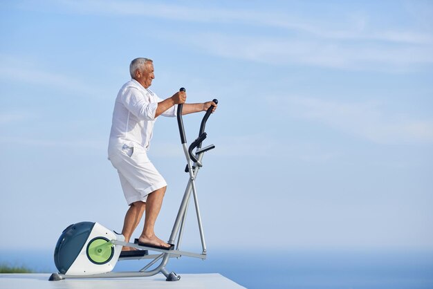 Gesunder älterer Mann, der auf dem Laufband im Fitnessstudio auf der modernen Terrasse mit Meerblick trainiert