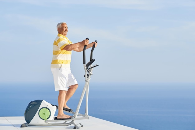 Gesunder älterer Mann, der auf dem Laufband im Fitnessstudio auf der modernen Terrasse mit Meerblick trainiert