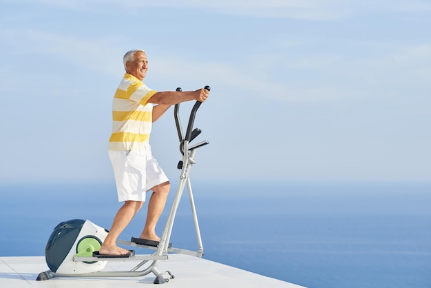 Gesunder älterer Mann, der auf dem Laufband im Fitnessstudio auf der modernen Terrasse mit Meerblick trainiert