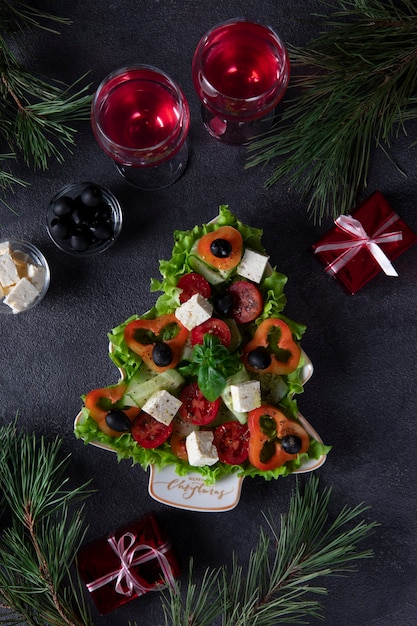 Gesunder griechischer salat in teller serviert als weihnachtsbaum mit festlicher dekoration und zwei gläser wein auf dunklem hintergrund. vertikales format. ansicht von oben