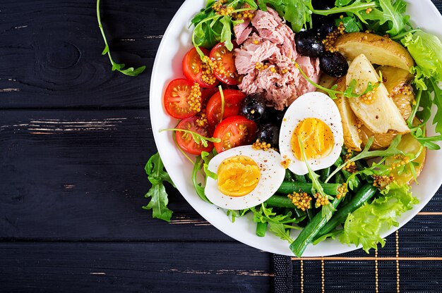 Gesunder herzhafter Salat des Thunfischs, der grünen Bohnen, der Tomaten, der Eier, der Kartoffeln, der Nahaufnahme der schwarzen Oliven in einer Schüssel auf dem Tisch. Salat Nicoise. Französische Küche. Ansicht von oben. Flach liegen