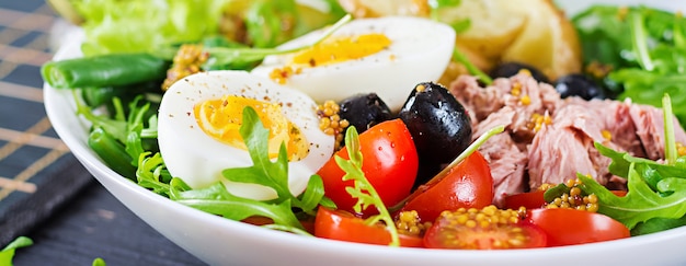 Foto gesunder herzhafter salat des thunfischs, der grünen bohnen, der tomaten, der eier, der kartoffeln, der nahaufnahme der schwarzen oliven in einer schüssel auf dem tisch. salat nicoise. französische küche. banner