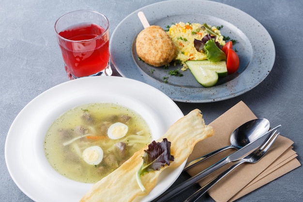 Gesunder Lebensmittelhintergrund, Restaurantgeschäftsessen. Nudelsuppe, Gemüserisotto mit Hähnchenschnitzel, erfrischendes Beerengetränk auf grauem Tisch, Besteck und leere Visitenkarte für Kopierraum