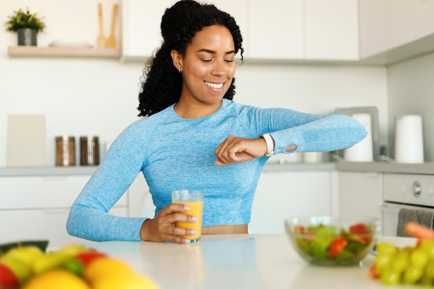 Gesunder Lebensstil Fröhliche, fitte Afroamerikanerin im Sport-BH mit frischem Saft und Blick auf die Smartwatch am Handgelenk