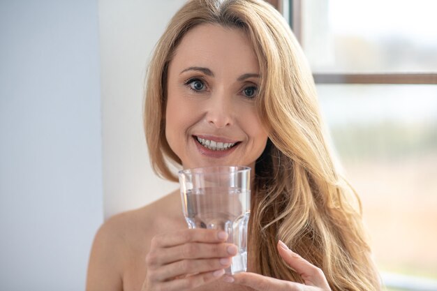 Gesunder Lebensstil. Glückliche schöne langhaarige Frau mit charmantem Lächeln mit nackten Schultern mit Glas Wasser am Morgen im Zimmer