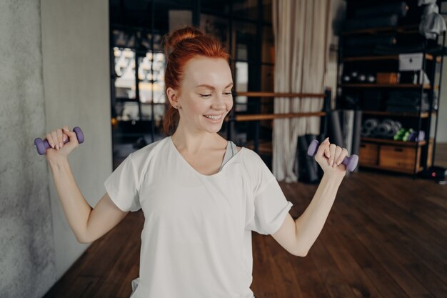 Gesunder Lebensstil. Junge, glückliche, schlanke, schöne Frau mit roten Haaren im Brötchen, die im Fitnessstudio trainiert, Hanteln hält und für starke Arme trainiert, von der Kamera wegschaut und lächelt