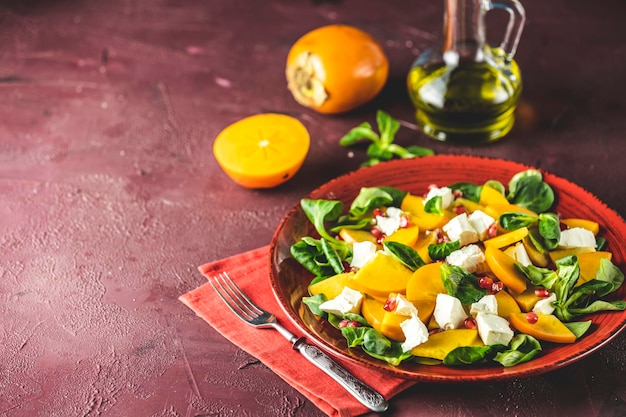 Gesunder Salat mit Kaki-Doucette, Feldsalat, Feldsalat und Feta-Käse auf einem roten Teller auf rotem Hintergrund. Superfoods Vitamin Herbst- oder Winter-Kakisalat