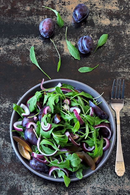 Gesunder Salat mit Rucola, Pflaume und blauen Zwiebeln in einer Schüssel auf einem stilvollen schäbigen Hintergrund