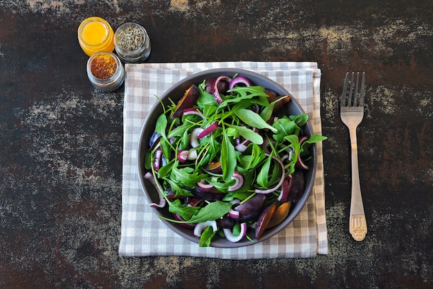 Gesunder Salat mit Rucola, Pflaume und blauen Zwiebeln in einer Schüssel auf einem stilvollen schäbigen Hintergrund