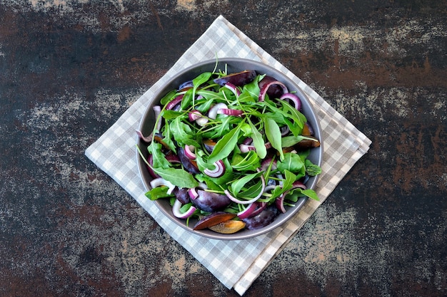 Gesunder Salat mit Rucola, Pflaume und blauen Zwiebeln in einer Schüssel auf einem stilvollen schäbigen Hintergrund