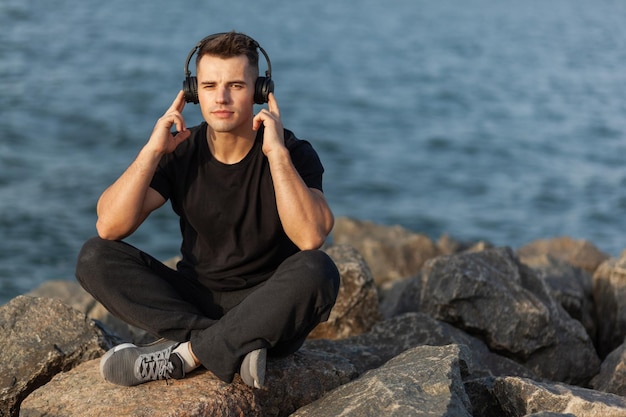 Gesunder Sportler, der Musik hört, während er auf Steinen an der Küste sitzt