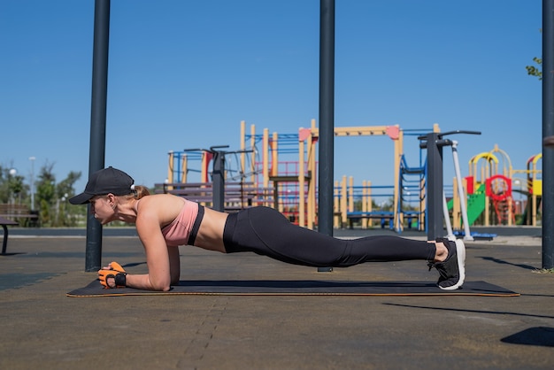 Gesunder und aktiver Lebensstil. Sport und Fitness. Glückliche Frau in Sportkleidung, die an sonnigen Sommertagen auf dem Sportplatz trainiert und ihre Bauchmuskeln in Plankenposition trainiert