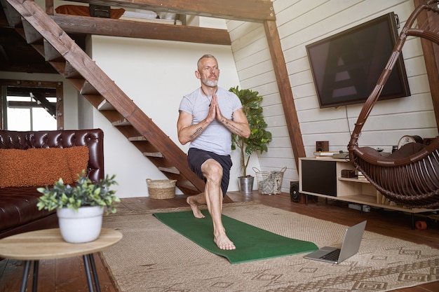 Gesunder und ruhiger Mann mittleren Alters, der zu Hause Yoga praktiziert, während er sich ein Online-Video-Tutorial anschaut