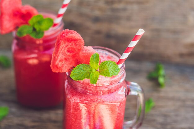 Gesunder Wassermelonen-Smoothie in Einmachgläsern mit Minze und gestreiften Strohhalmen auf Holzhintergrund.