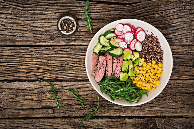 Gesundes Abendessen. Buddha-Schüsselmittagessen mit gegrilltem Rindfleischsteak und Quinoa, Mais, Avocado, Gurke und Arugula auf Holztisch. Fleischsalat. Flach liegen. Ansicht von oben