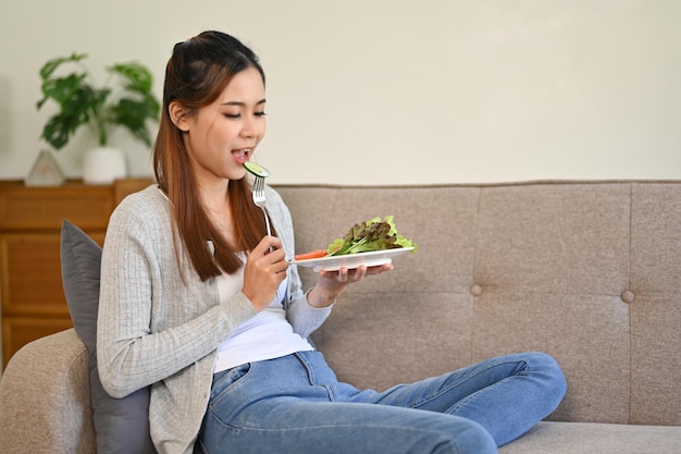 Gesundes asiatisches Mädchen, das ihren gesunden Salat isst, während es sich in ihrem Wohnzimmer entspannt