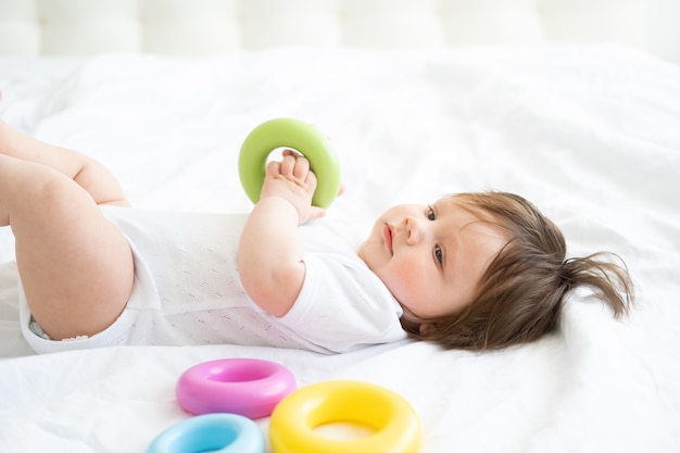 Gesundes Baby, das auf Bett liegt und mit buntem Plastikkreisspielzeug spielt.