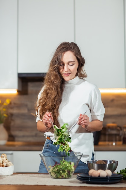 Gesundes Essen - Gemüsesalat. Diät. Diät-Konzept. Junge lockige Frau, die Gemüsesalat in ihrer Küche vorbereitet. Gesundes Lebensstilkonzept, schönes lächelndes Mischgemischgemüse der Frau.