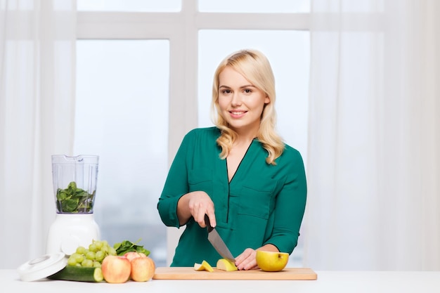 Gesundes Essen, Kochen, vegetarisches Essen, Ernährung und People-Konzept - lächelnde junge Frau mit Mixer und Messer, die zu Hause Obst und Gemüse auf dem Schneidebrett hackt