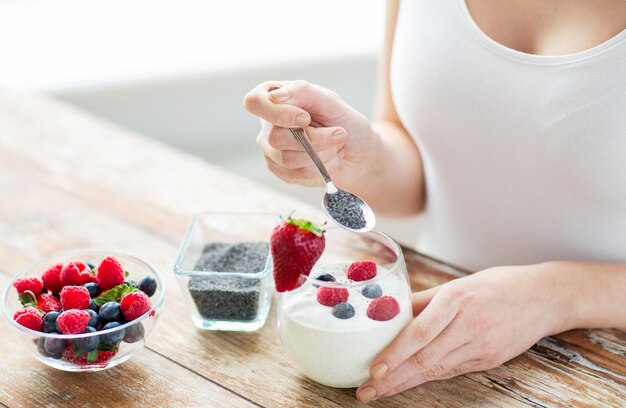 Gesundes Essen, vegetarisches Essen, Ernährung und People-Konzept - Nahaufnahme von Frauenhänden mit Joghurt, Beeren und Mohn- oder Chiasamen auf Löffel