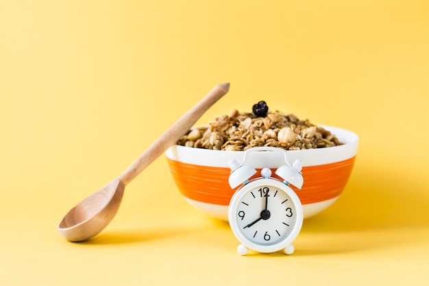 Gesundes Essen. Wecker vor einem gebackenen Müsli aus Hafer, Nüssen und Rosinen in einer Schüssel und einem Holzlöffel auf einer gelben Oberfläche