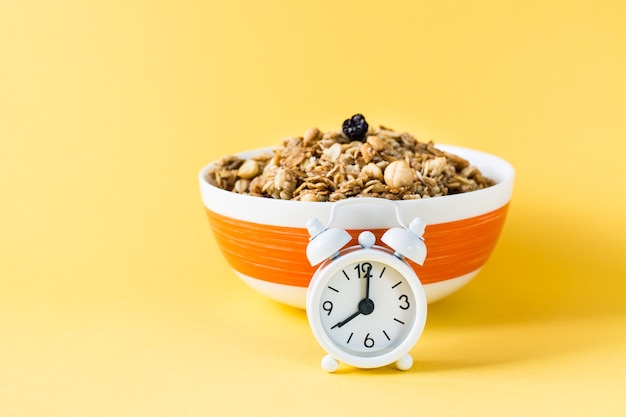 Gesundes Essen. Wecker vor gebackenem Müsli aus Hafer, Nüssen und Rosinen in einer Schüssel auf gelber Oberfläche