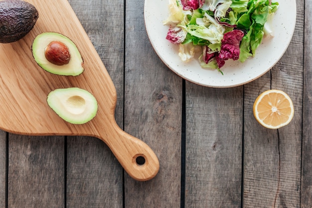 Gesundes Esskonzept flach lag. Mediterrane Ernährung, Teller mit frischen grünen Salatblättern, lila Radicchio-Blättern, Schneidebrett mit Avocadohälften, Kern, Zitrone auf Holztisch