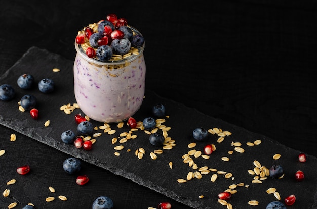 Gesundes Fitnessfutter. Glas selbst gemachter Joghurt mit Blaubeeren, Granatapfelsamen und Hafer auf Schwarzem. Vorderansicht, Exemplar