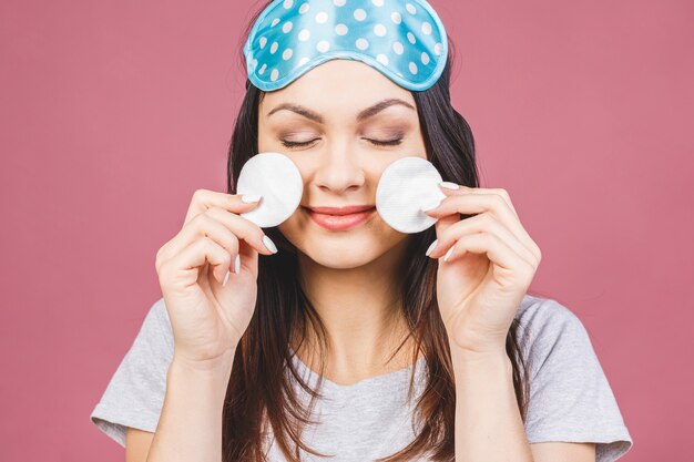 Gesundes frisches Mädchen, das Make-up von ihrem Gesicht mit Wattepad entfernt. Schönheitsfrau, die ihr Gesicht mit Wattestäbchenauflage lokalisiert auf rosa Hintergrund reinigt. Hautpflege- und Schönheitskonzept. Schlafmaske.