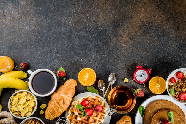 Gesundes Frühstück, das Konzept, verschiedenes Morgenlebensmittel - Pfannkuchen, Waffeln, Hörnchenhafermehlsandwich und Granola mit Jogurt, Frucht, Beeren, Kaffee, Tee, Orangensafthintergrund isst