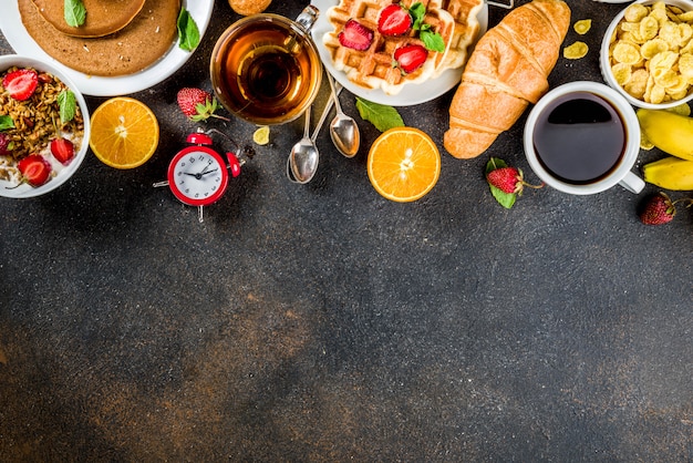 Gesundes Frühstück, das Konzept, verschiedenes Morgenlebensmittel - Pfannkuchen, Waffeln, Hörnchenhafermehlsandwich und Granola mit Jogurt, Frucht, Beeren, Kaffee, Tee, Orangensafthintergrund isst