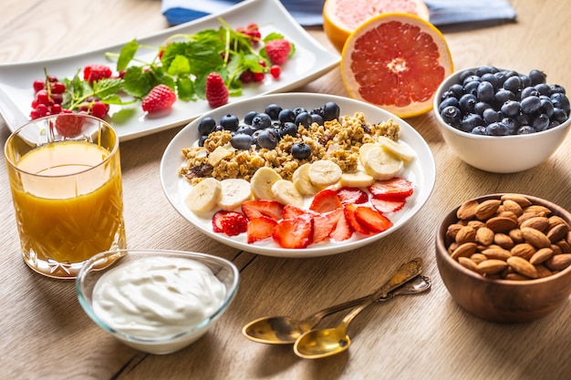 Foto gesundes frühstück serviert mit einem teller mit joghurt-müsli-heidelbeeren, erdbeeren und banane. morgentisch müsli mandeln beeren zitrusfrüchte saft und grüne kräuter.