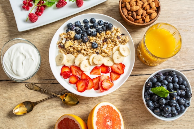 Gesundes Frühstück serviert mit einem Teller mit Joghurt-Müsli-Heidelbeeren, Erdbeeren und Banane. Morgentisch Müsli Mandeln Beeren Zitrusfrüchte Saft und grüne Kräuter.