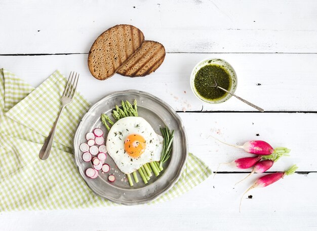 Gesundes Frühstücksset. Spiegelei mit Spargel, Rettichen, grüner Soße und Brot auf Weinlesemetallplatte über weißem Holztisch