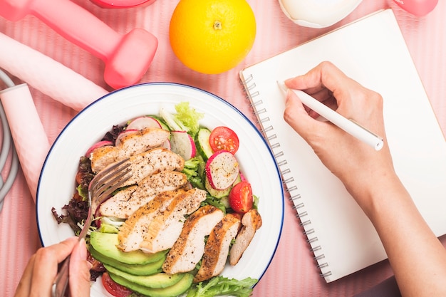 Gesundes Konzept mit Ernährungsnahrung in der Brotdose und Fitnessausrüstungen mit Frau, die Zeit schreibt, um auf Tagebuchbuch gesund zu werden