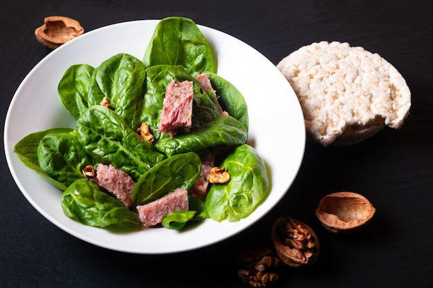 Gesundes Lebensmittelkonzept Spinatsalat mit französischem Presskopf in weißer Keramikschale auf schwarzem Schiefersteinbrett mit Kopienraum