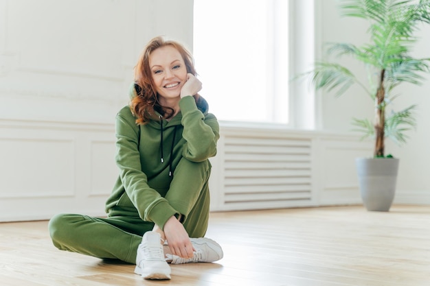 Gesundes Lifestyle-Konzept Schöne schlanke junge Frau mit rotem, welligem Haar hält die Hand unter dem Kinn, lächelt glücklich in Sportbekleidung gekleidet und hat einen flexiblen Körper, der auf dem Boden sitzt. Frau hat Heimtraining